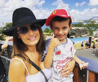 Woman in sunglasses and black hat smiles, holding a boy in a red cap and Liverpool FC shirt at a zoo with skyline in background.