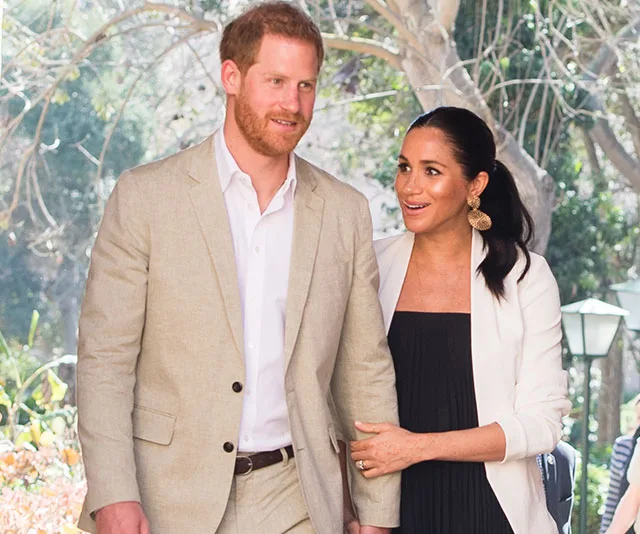 A couple smiling and walking outdoors, surrounded by greenery, wearing light-colored formal attire.