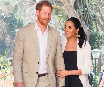A couple smiling and walking outdoors, surrounded by greenery, wearing light-colored formal attire.