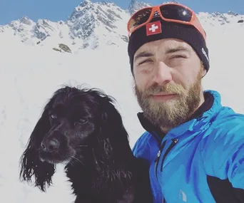 Man in winter attire with black dog in snowy mountain setting.