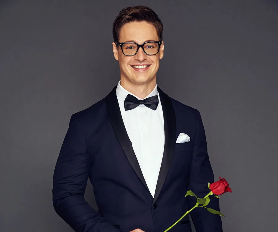 Man in a tuxedo holding a rose, wearing glasses, smiling at the camera against a gray background.