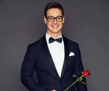 Man in a tuxedo holding a rose, wearing glasses, smiling at the camera against a gray background.
