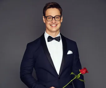Man in a tuxedo holding a rose, wearing glasses, smiling at the camera against a gray background.