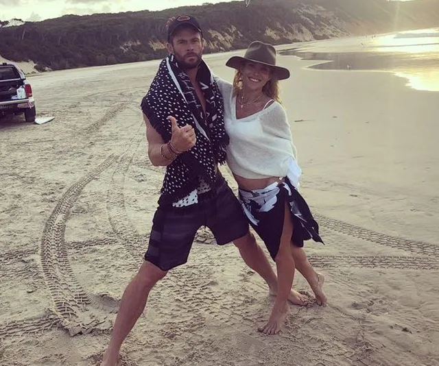 A couple posing playfully on a sandy beach with a vehicle and tire tracks in the background.