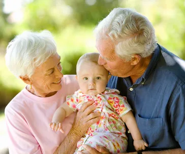 Elderly couple smiling and holding a baby, with the man gently kissing the baby's cheek outdoors.