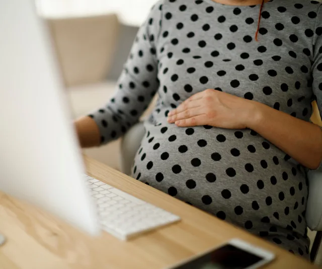 Pregnant woman in polka dot dress uses a desktop computer, hand on belly, sitting at a wooden desk in home office.