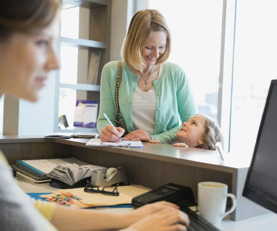 "Smiling woman and child at medical reception desk, filling out forms, with receptionist blurred in foreground."