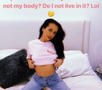 A woman in a white shirt and jeans, sits on a bed with a thought bubble emoji above her.