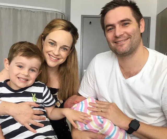 A family of four, including a young boy, woman, man, and newborn wrapped in a striped blanket, smiling in a room.