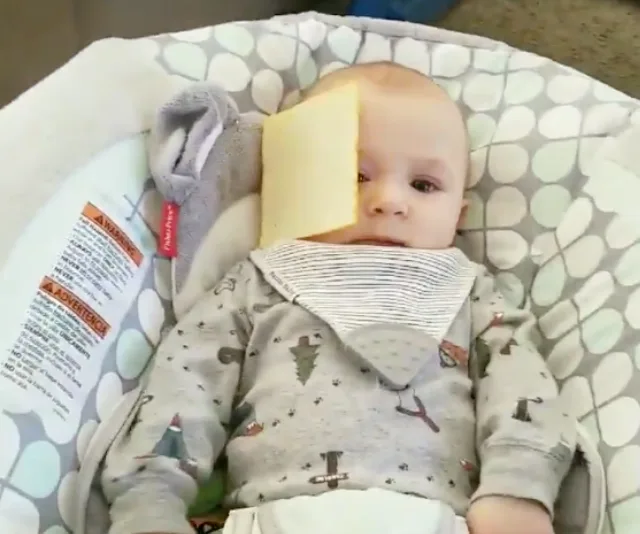 A baby wearing a patterned onesie is lying in a baby seat with a slice of cheese on their face.