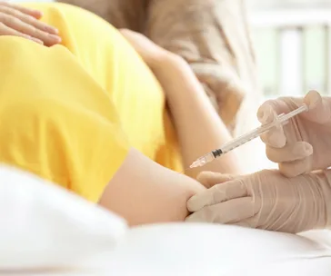A pregnant woman receives a vaccination in her arm from a healthcare professional wearing gloves.