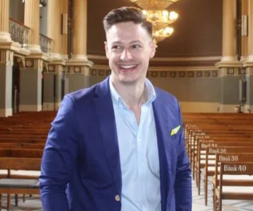 A man in a blue blazer smiles in a well-lit, elegant hall with wooden benches marked with numbers.
