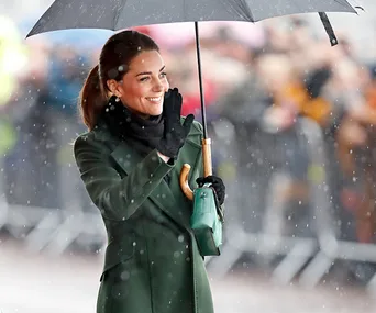 A woman in a green coat holds an umbrella and waves in the rain, with blurred onlookers in the background.