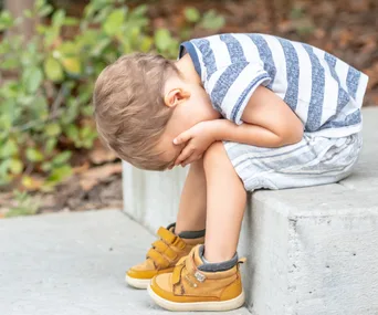 A young child sits on a concrete step, head in hands, appearing upset or sad, wearing a striped shirt and yellow shoes.