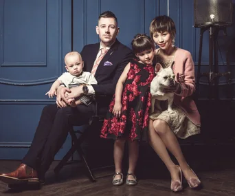 A family of four and a dog pose in front of a blue wall; the mother holds a small white dog.