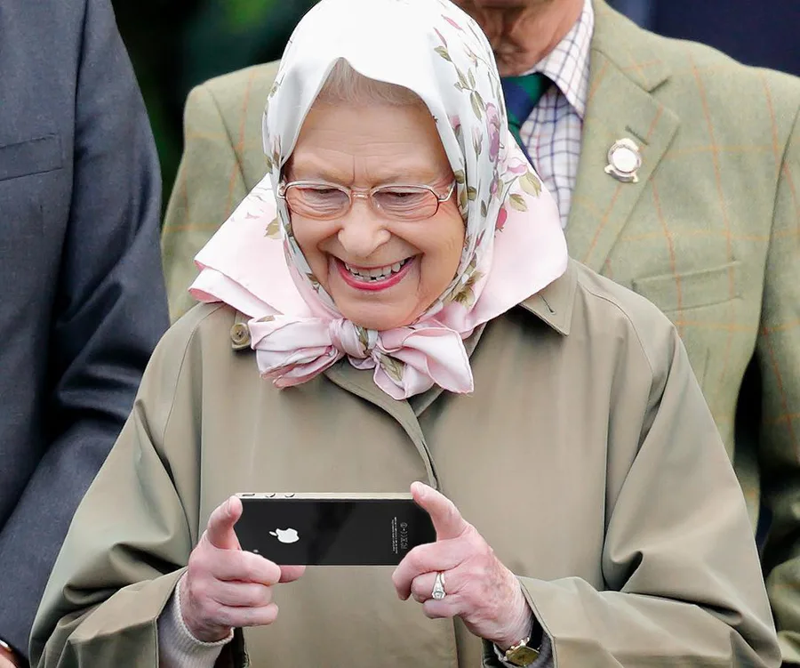 Elderly woman in headscarf and coat smiles while using a smartphone outdoors.