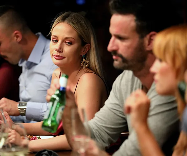 A woman looks at a man holding a beer bottle at a dinner table, with two others in the background.
