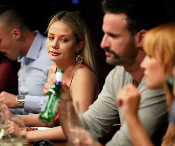 A woman looks at a man holding a beer bottle at a dinner table, with two others in the background.