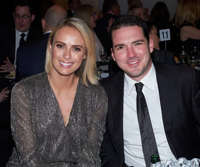 A smiling woman in a sparkly dress and a man in a suit sit together at an event, surrounded by people.