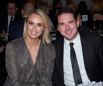 A smiling woman in a sparkly dress and a man in a suit sit together at an event, surrounded by people.