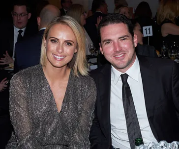 A smiling woman in a sparkly dress and a man in a suit sit together at an event, surrounded by people.