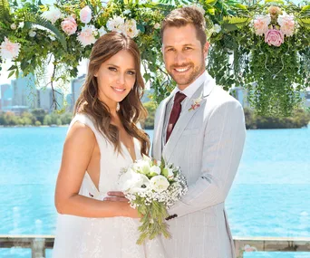 Elly and Mark from Neighbours smiling at their wedding ceremony by a lake.