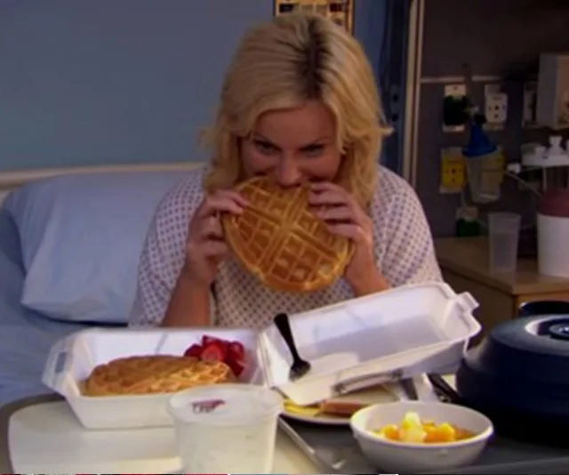 A person in a hospital bed eagerly biting into a waffle with food containers and fruit on the tray.