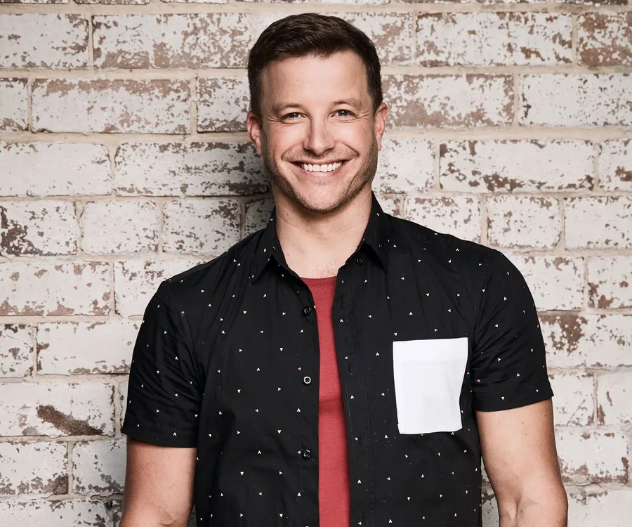 Man smiling in front of a brick wall, wearing a black shirt with small patterns and a red t-shirt underneath.
