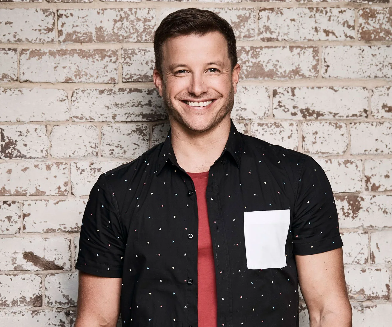 Man smiling in front of a brick wall, wearing a black shirt with small patterns and a red t-shirt underneath.
