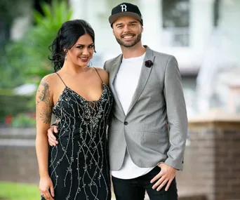 Man in grey blazer and cap with woman in black dress posing together, outdoors with greenery and blurred building in background.