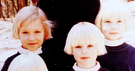 Children with blonde hair wearing dark clothing, standing together outdoors, with snow visible in the background.