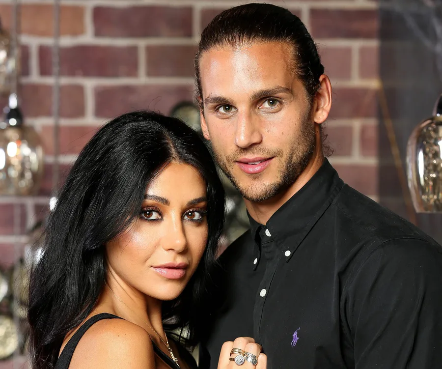 A couple poses together, both with dark hair; the woman has long hair, the man wears a black shirt, brick wall background.