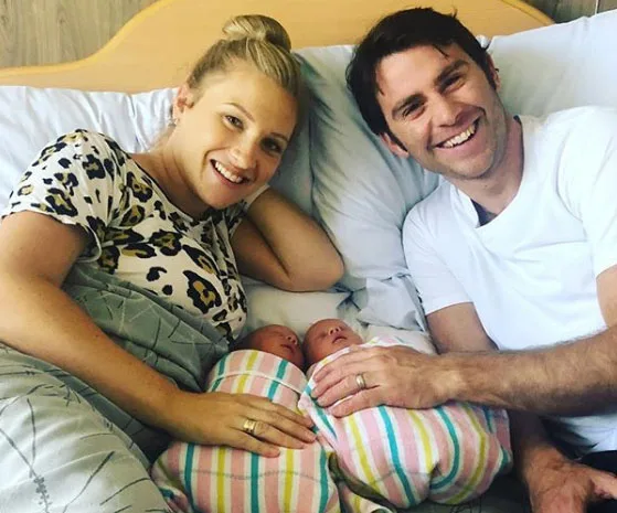 Happy couple lying in bed with twin newborns, swaddled in colorful blankets, smiling at the camera.