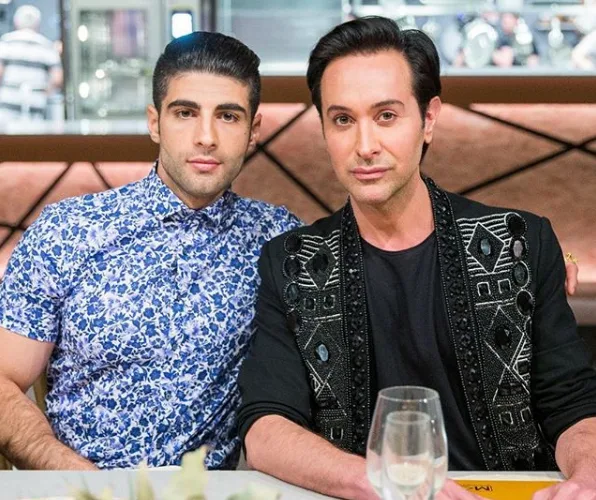 Two men sitting together at a dining table, one in a blue floral shirt and the other in a black embellished jacket.
