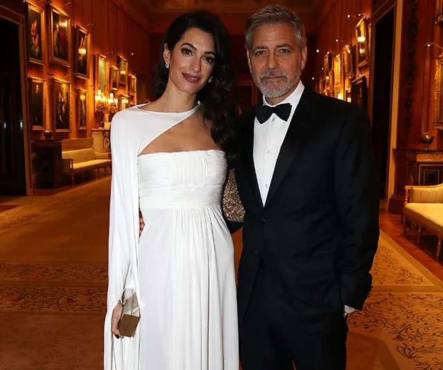 Couple elegantly dressed at Buckingham Palace event, posing in opulent room with portraits and soft lighting.