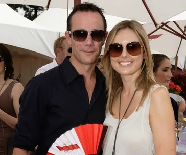 Couple wearing sunglasses, smiling outdoors under umbrellas at an event; man holds a fan.