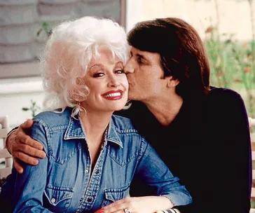 Man kisses smiling woman in denim shirt with white hair.