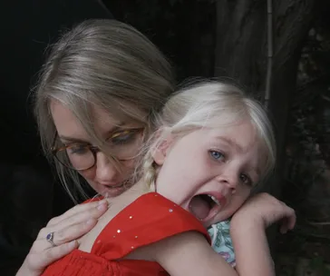 A woman with glasses cuddles a blonde child in a red dress, the child looking surprised or distressed.