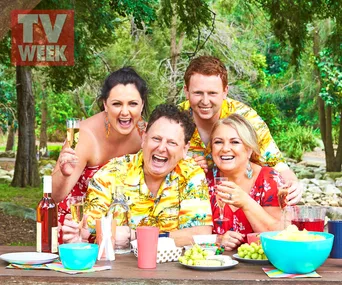 Four people in colorful clothes enjoying a picnic outdoors, laughing and holding drinks, surrounded by trees and snacks.