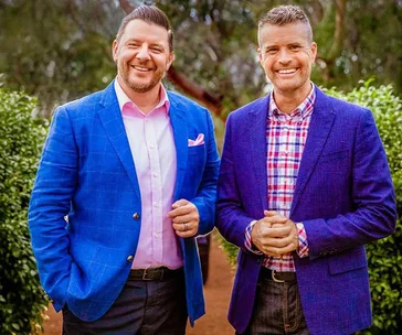 Two men in blue blazers smiling outdoors, with trees and greenery in the background.