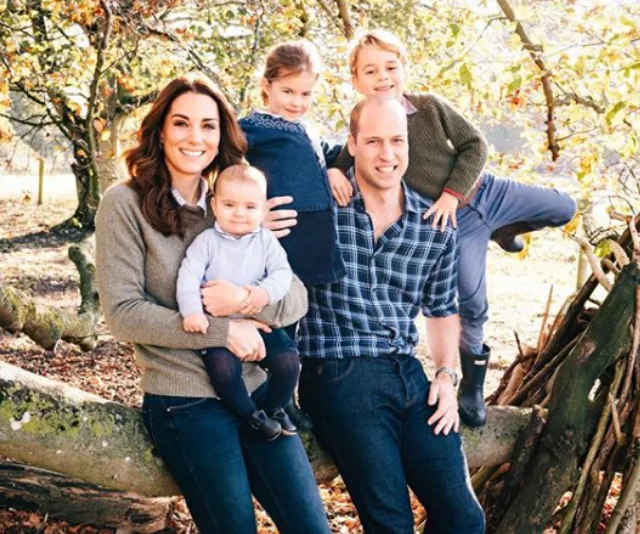 A family of five poses outdoors in front of autumn trees, smiling and wearing casual clothing.
