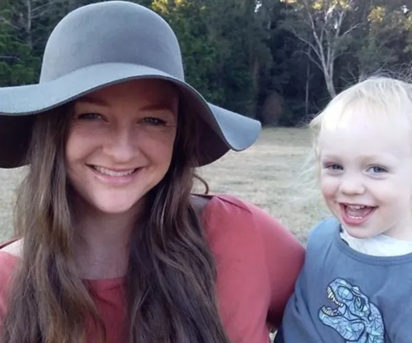 A smiling woman in a hat holds a happy toddler in a grassy field with trees in the background.