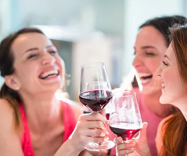 Three women smiling and clinking glasses of red wine in a celebratory setting.