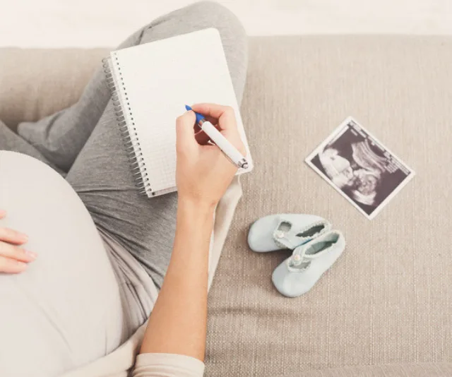 Pregnant woman writing in notebook with sonogram and baby shoes nearby.