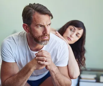 Man sitting on edge of bed, looking pensive, while woman behind him rests her head on his shoulder, appearing concerned.