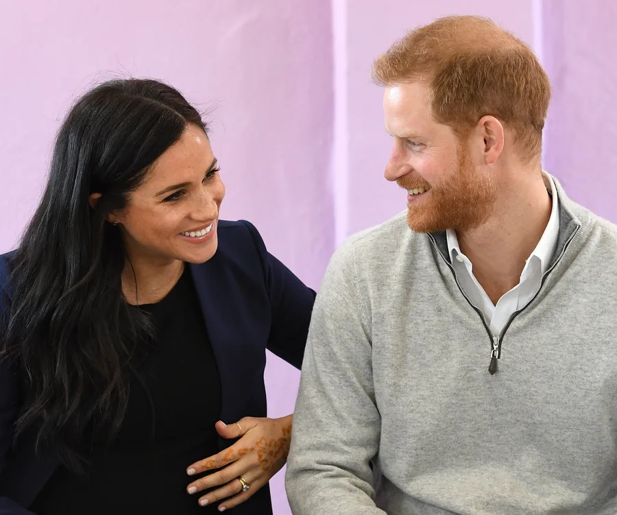 Two people smiling at each other; one with dark hair, the other with red hair, seated against a purple wall.