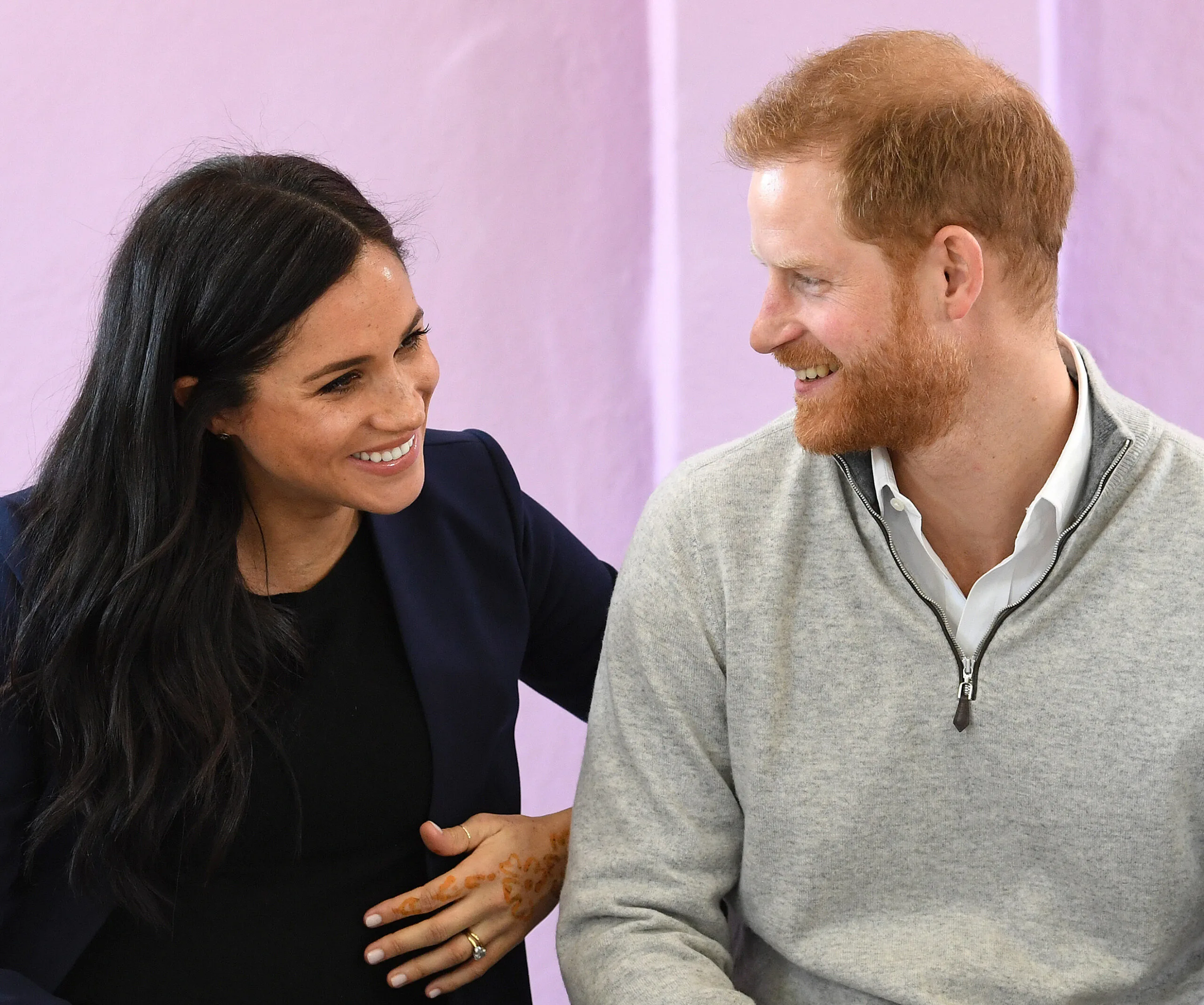 Two people smiling at each other; one with dark hair, the other with red hair, seated against a purple wall.
