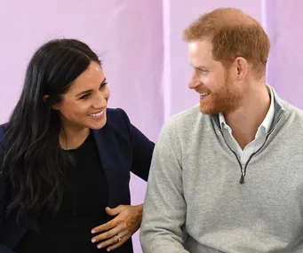 Two people smiling at each other; one with dark hair, the other with red hair, seated against a purple wall.