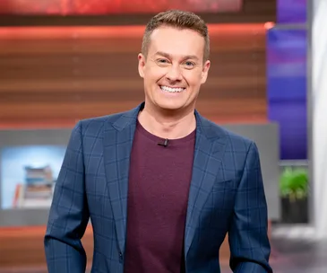 A smiling TV host wearing a plaid blazer stands on a brightly lit studio set.