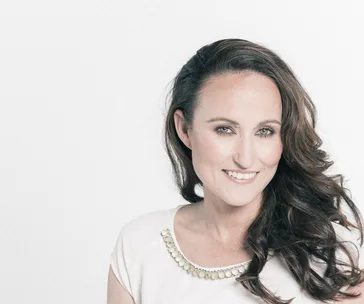 A woman with long brown hair, smiling and wearing a white top with decorative collar, against a plain background.